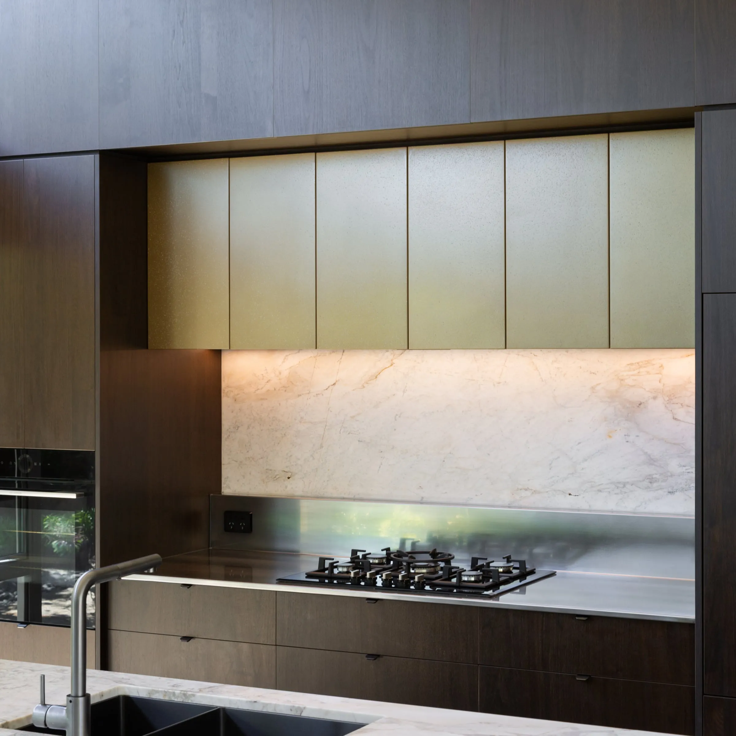 Cooking wall with brass uppers, lit stone splashback and stainless benchtop