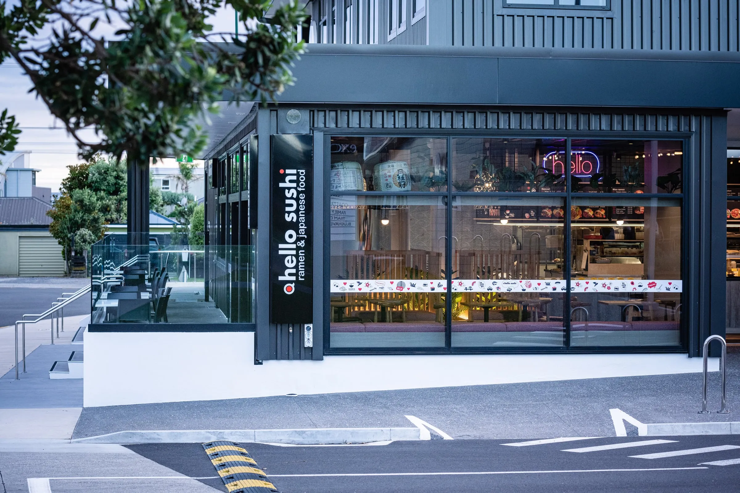 Dusk exterior of the black-clad Hello Sushi corner shopfront, neon glowing through glazing