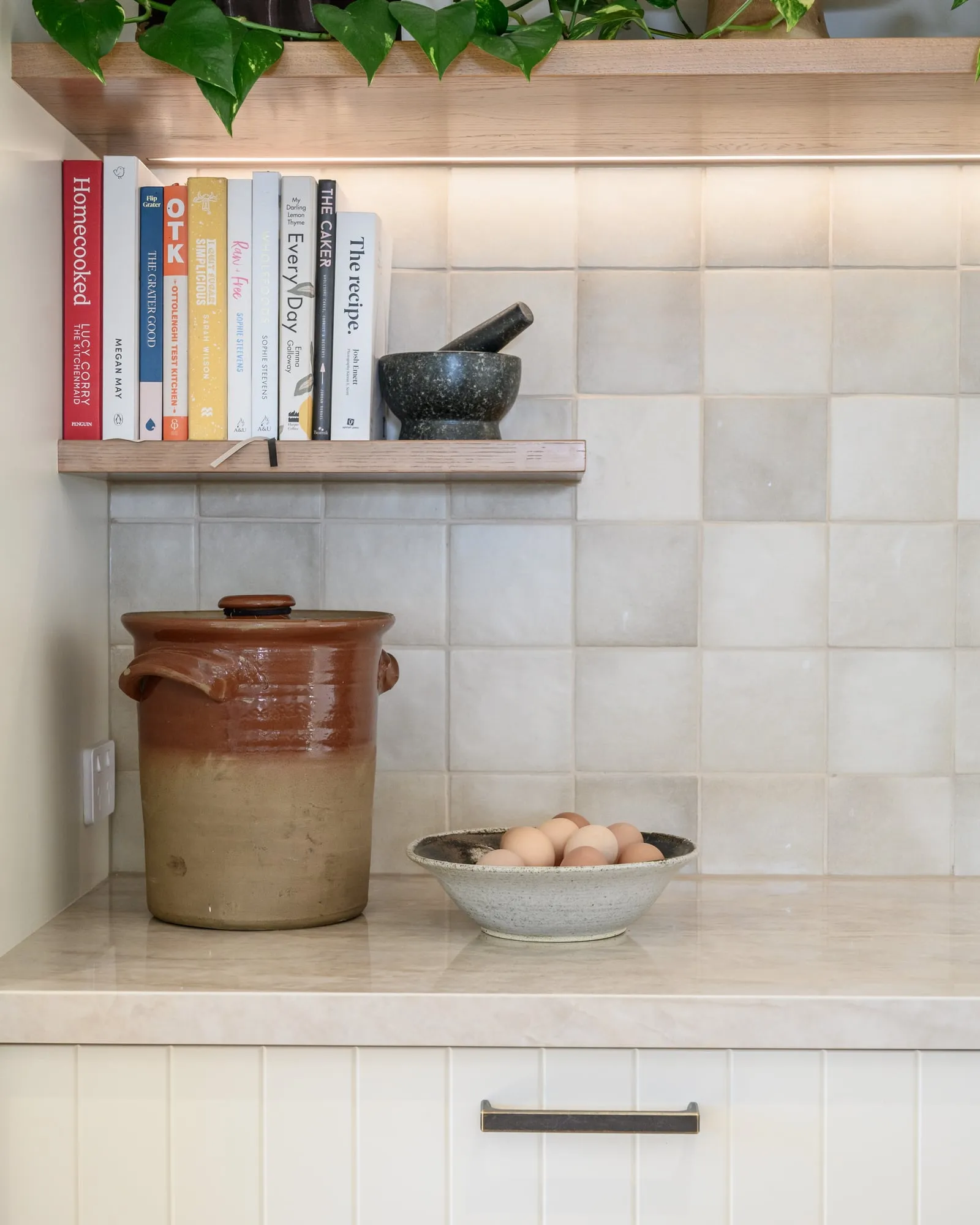 Oak floating shelf with cookbooks above a lit handmade-look tile splashback