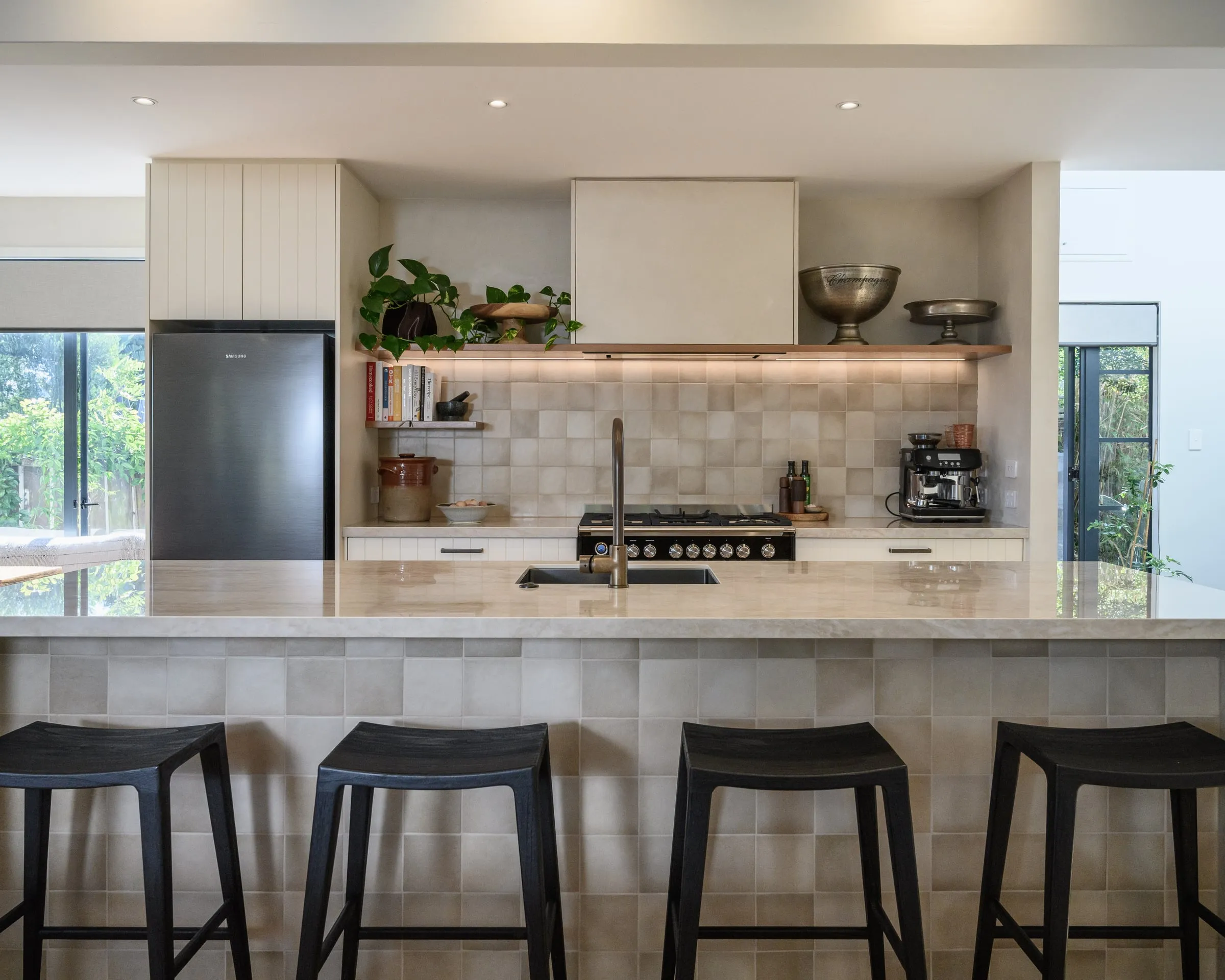 Cream panelled kitchen with a stone island, black stools and a tiled cooking wall