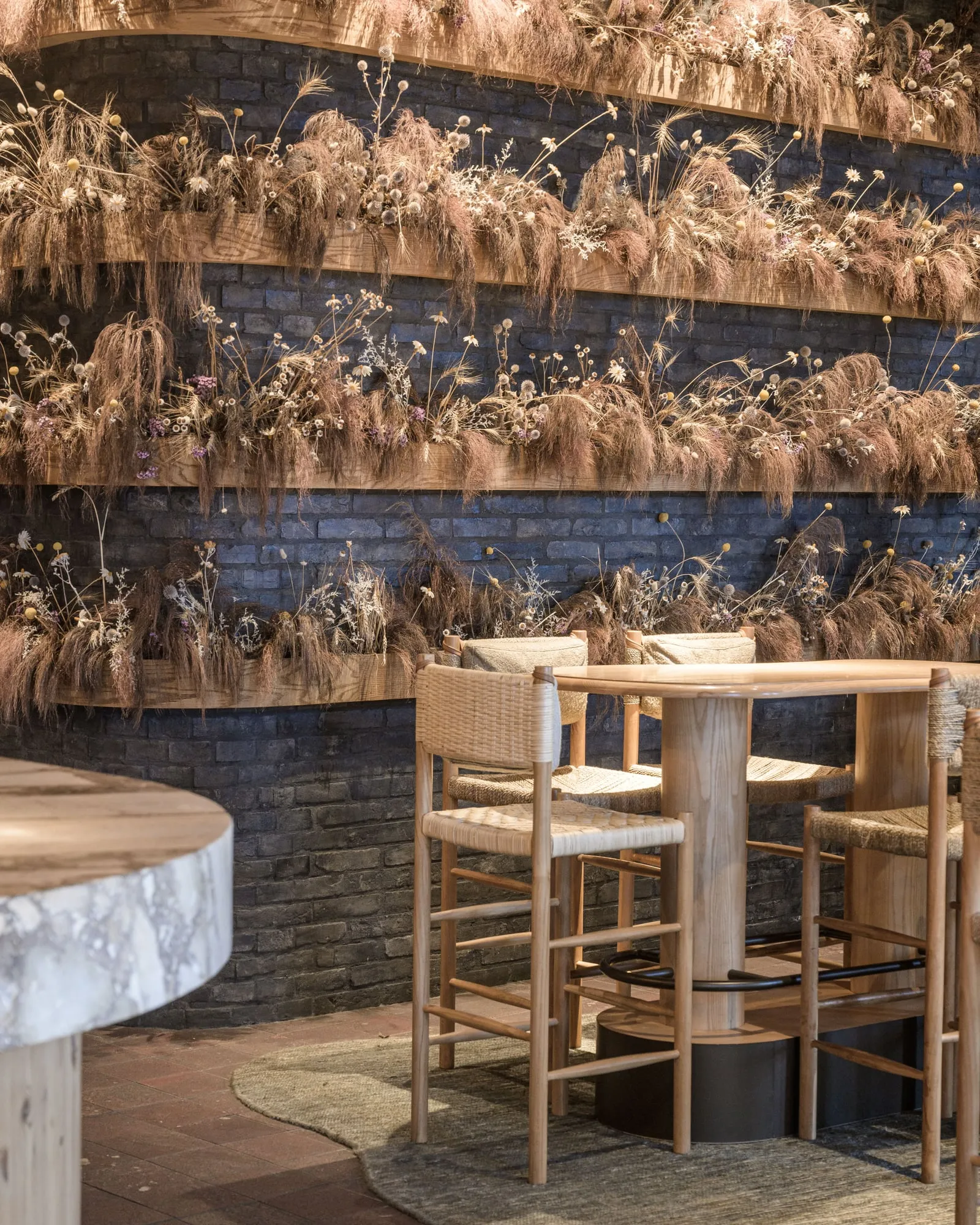 Dried-grass installation over dark brick with pale Ash high table and woven stools