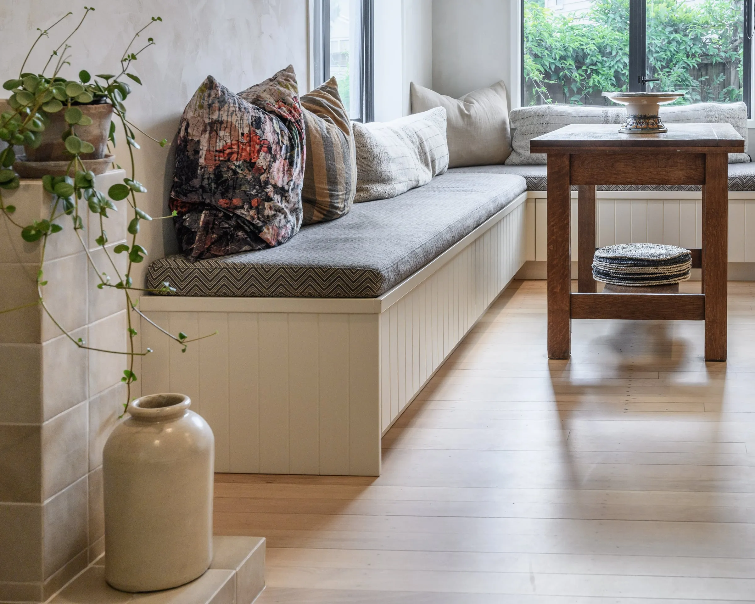 Panelled window seat with cushions and a vase beside full-height glazing